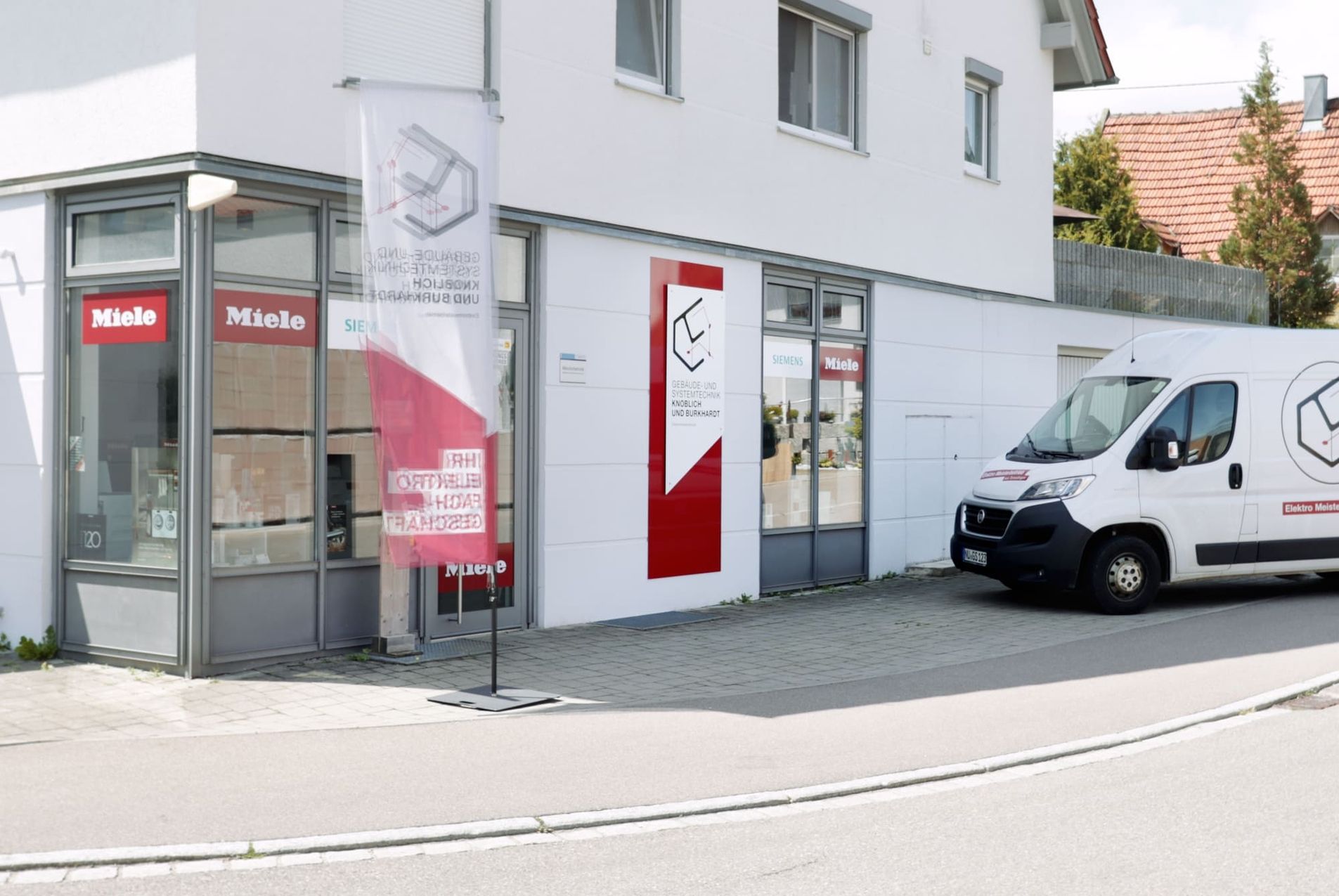 Außenansicht eines Geschäfts mit Ausstellungsstücken von Miele und Siemens, vor dem an einem sonnigen Tag ein Lieferwagen geparkt ist.